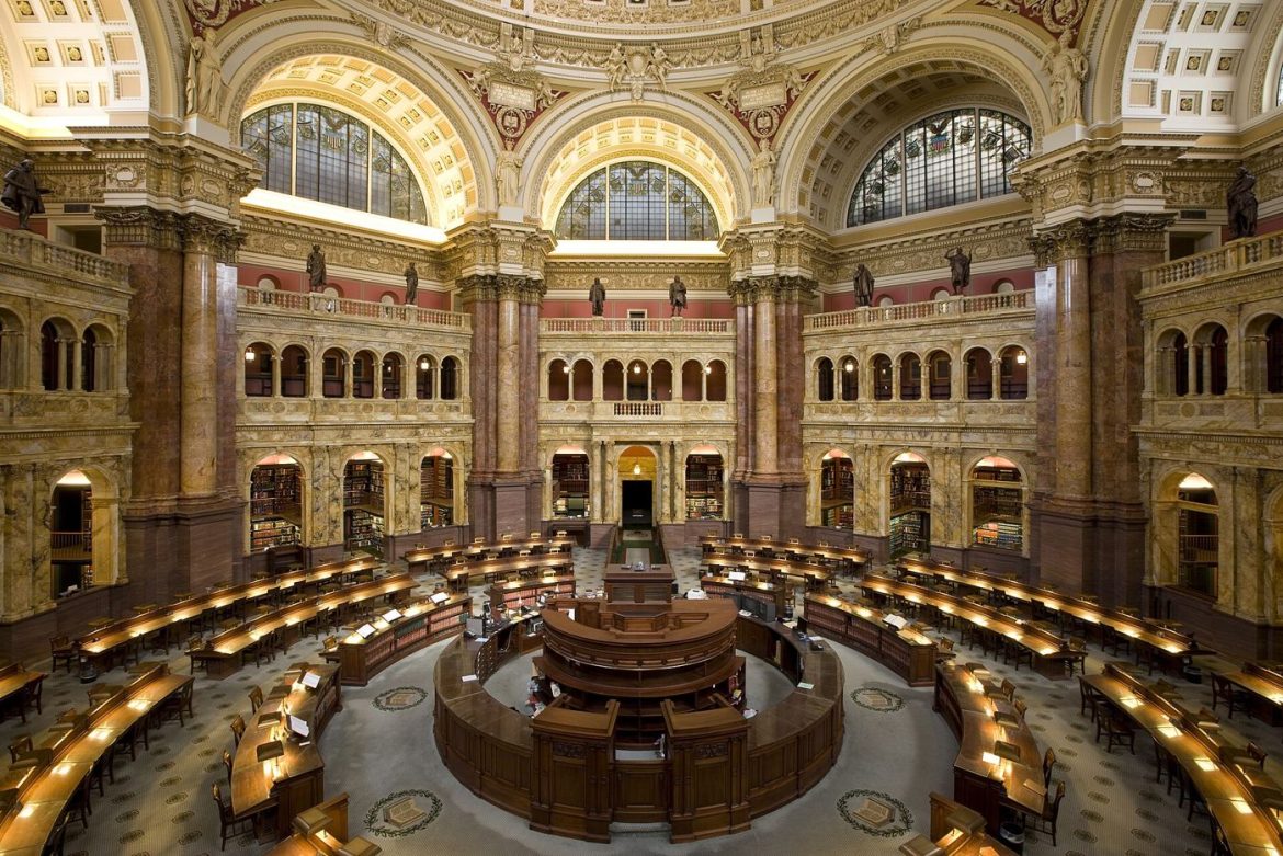 Readers study quietly at long wooden tables inside a bright public library reading room.
